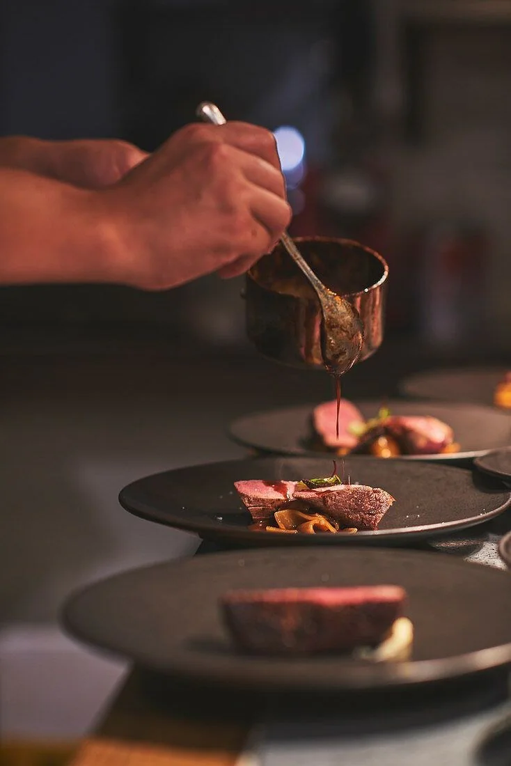 Mãos do chef regando com molho escuro os pratos de carne alinhados no passe