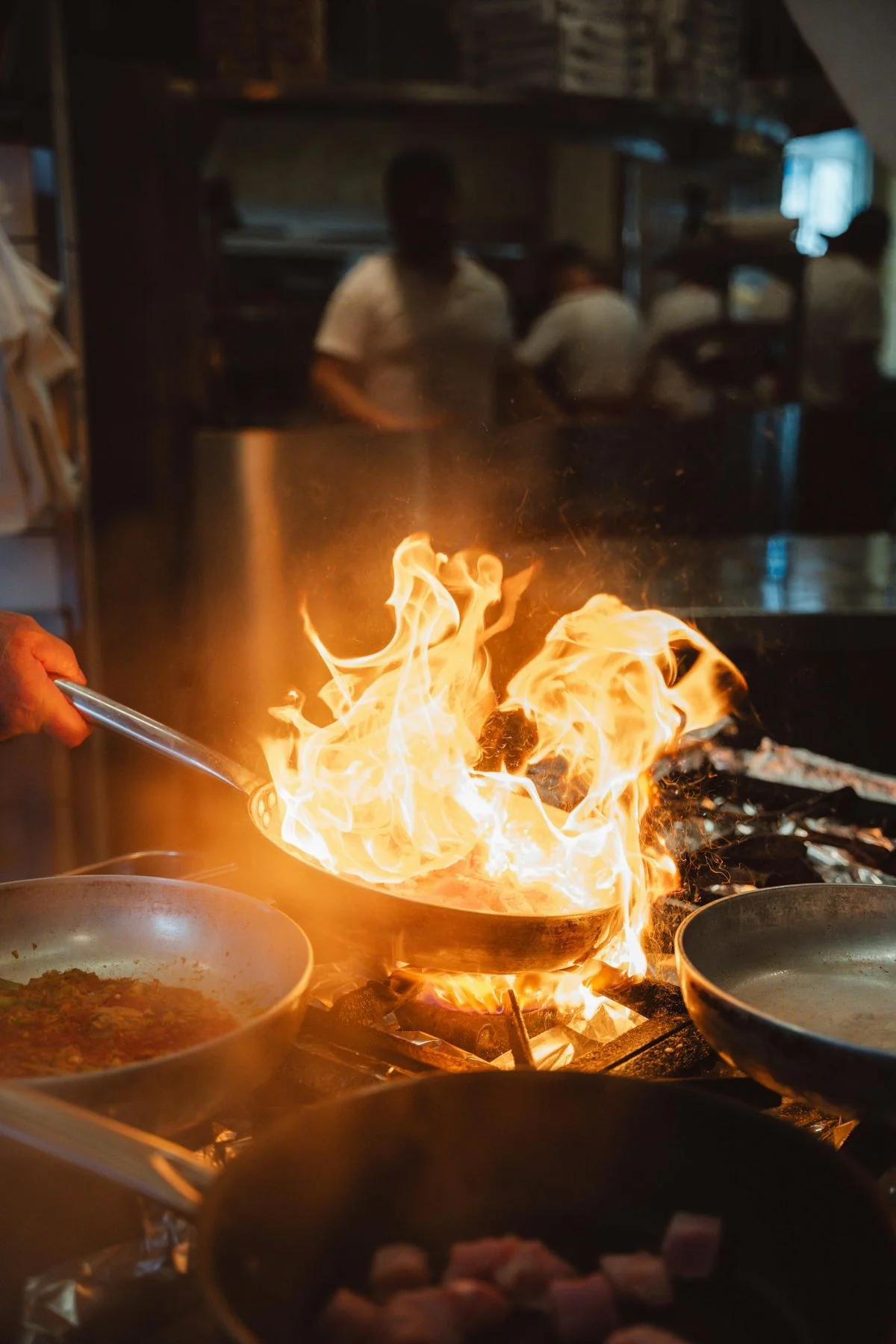 Frigideira em chamas sobre o fogão da cozinha, com a equipe desfocada ao fundo