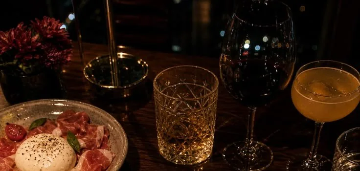 Mesa de jantar com burrata e presunto cru, flores, uma taça de vinho tinto e um coquetel